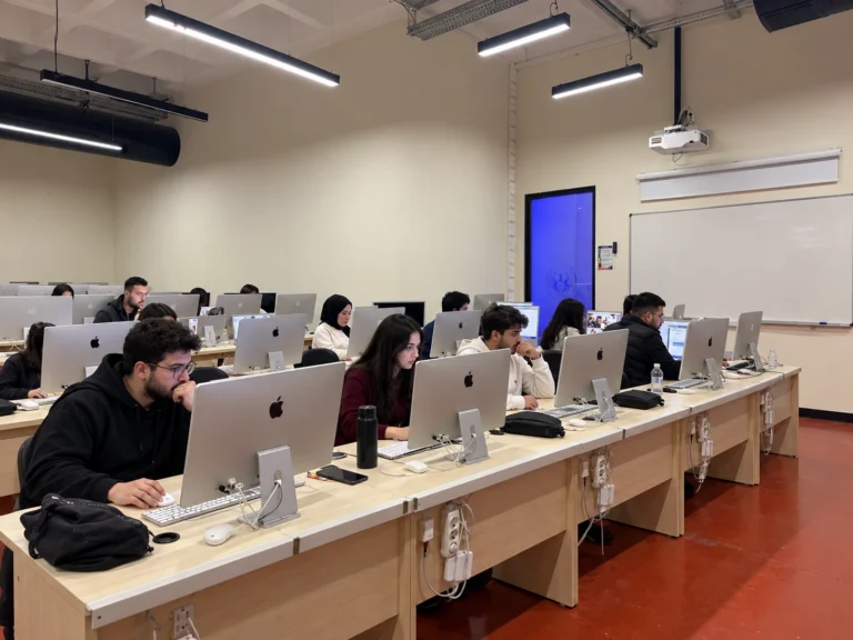 Mac bilgisayarların bulunduğu tasarım laboratuvarında, masaların iki yanında oturarak çalışan katılımcıların genel görünümü.